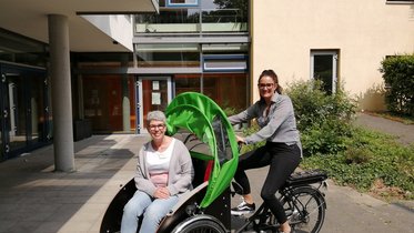Eine Frau sitzt auf einer Fahrradrikscha. Eine jüngere Frau sitzt auf dem Sattel.
