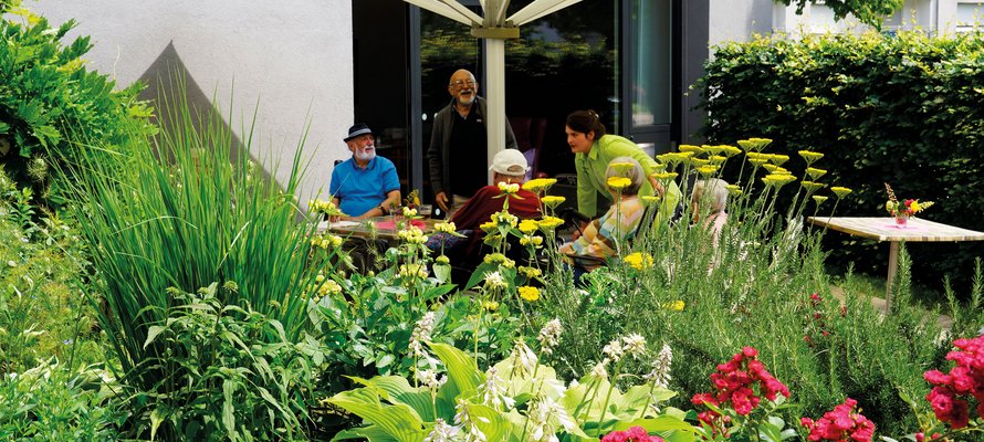 Blumenbeet, im Hintergrund Menschen, die unter einem Sonnenschirm sitzen.