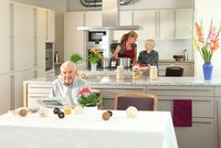 Senior sitzt an einem schön gedeckten Tisch, im Hintergrund die Wohnküche mit zwei Personen