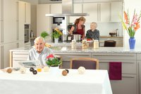 Senior sitzt an einem schön gedeckten Tisch, im Hintergrund die Wohnküche mit zwei Personen